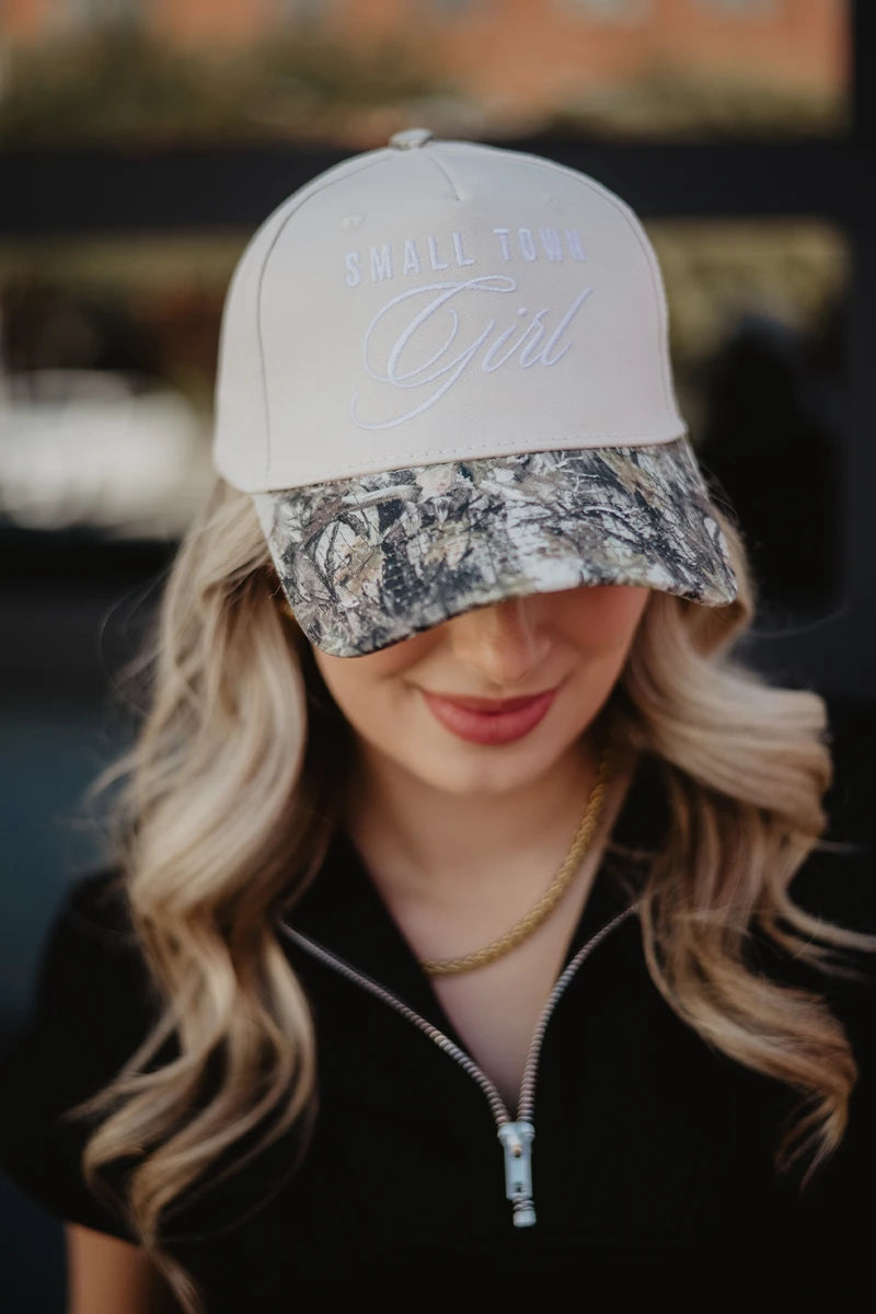 A woman with long, wavy blonde hair smiles while wearing a Katydid Small Town Girl Vintage Trucker Hat for Women with white lettering and a camo brim, paired with a black zip-up top.
