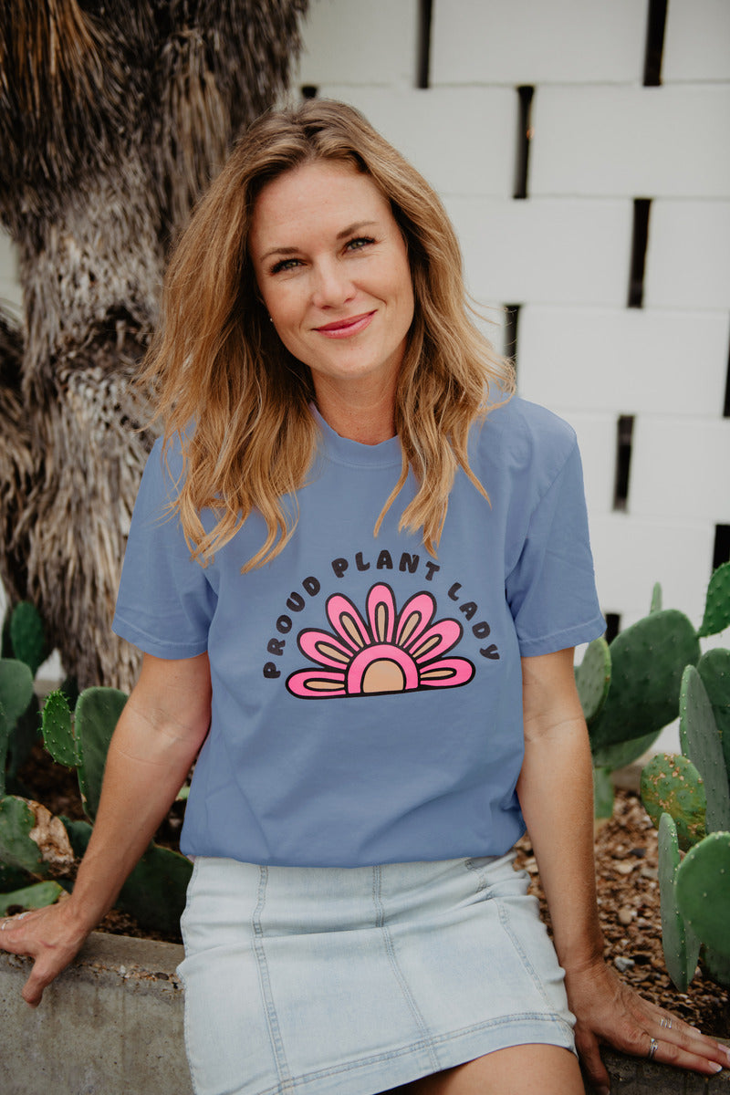 A woman with wavy blonde hair sits outdoors among cacti, smiling and wearing the Katydid Proud Plant Lady Graphic Tee for Women with a pink and yellow flower design, paired with a light denim skirt.