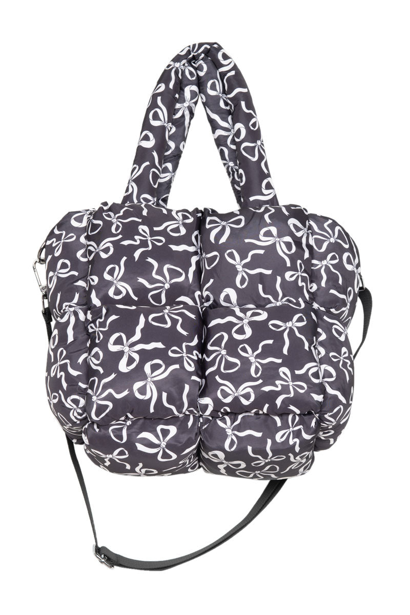 The Katydid Black Bow Print Puffer Tote is a quilted bag with a white abstract bow pattern, two short handles, and a detachable long shoulder strap.