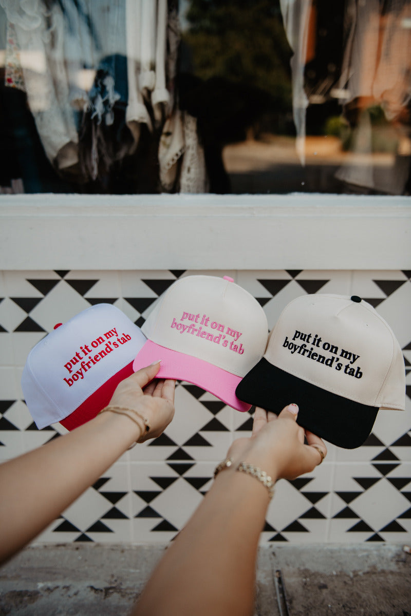Three hands hold trucker hats with the phrase "put it on my boyfriend's tab." The hats are white, pink, and beige with playful, casual vibes.