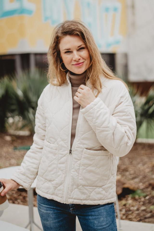 A woman with long blonde hair smiles at the camera outdoors, wearing the Katydid Cream Cute Garment Washed Quilted Jacket and jeans. She stands before green plants and a blue-and-yellow mural, creating a lively background.
