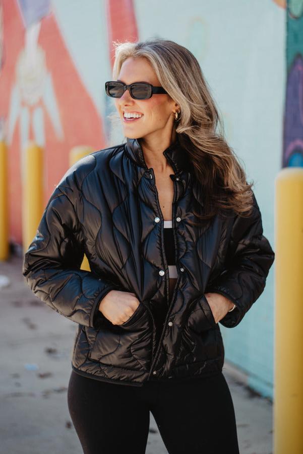 A woman with long, wavy hair and sunglasses stands outdoors wearing black pants and the Katydid Black Lightweight Buttoned Quilted Puffer Jacket. Her hands are in her pockets, with a colorful mural and yellow poles behind her.