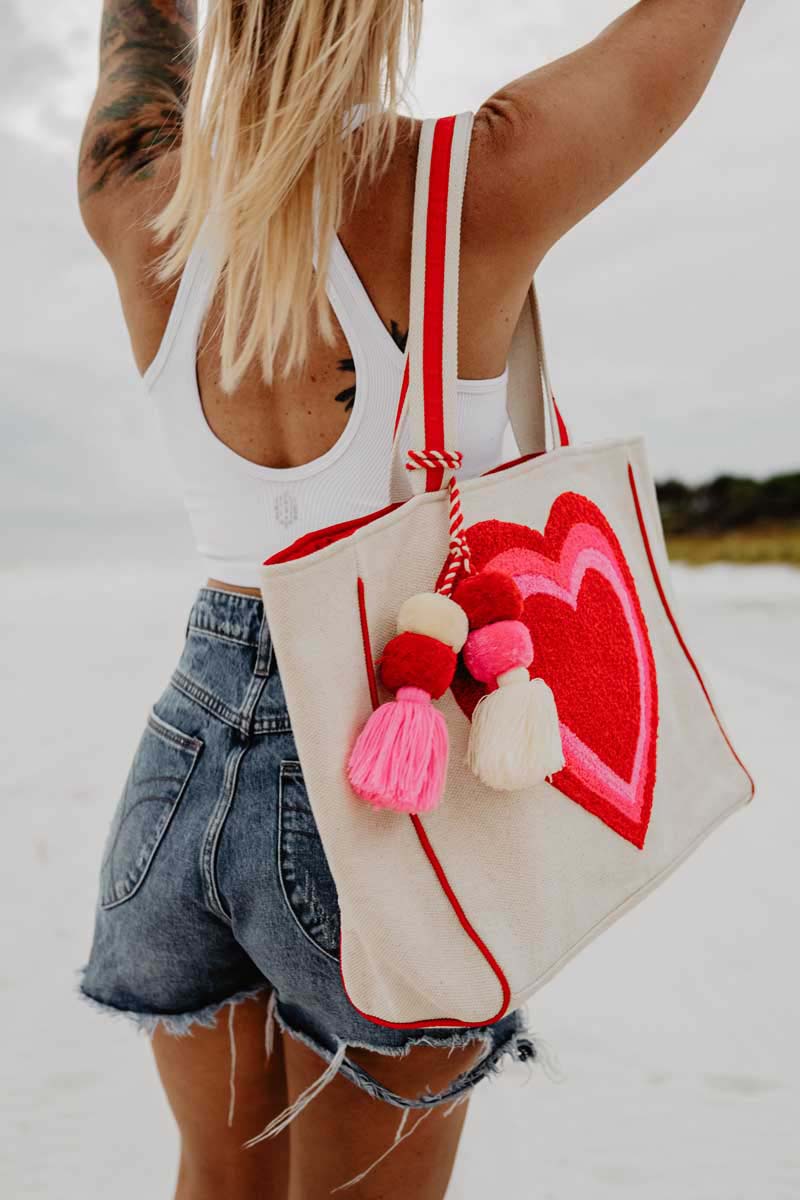 A woman with blonde hair in a white crop top and denim shorts stands on a beach, holding up a tote with red hearts and colorful tassels.