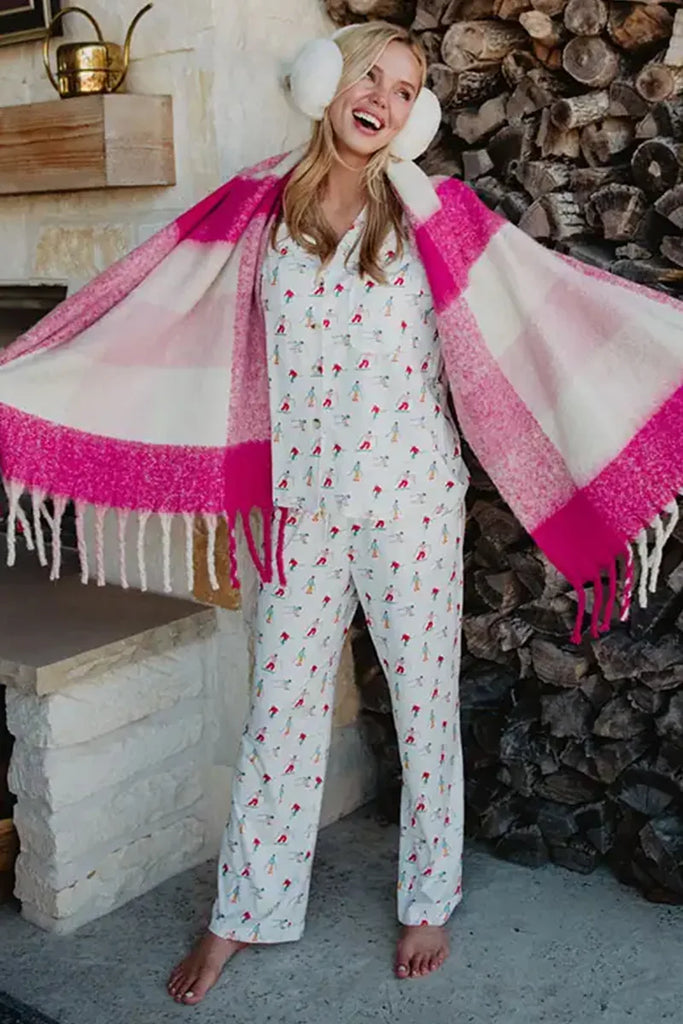 A woman wearing the Katydid Apres Ski Matching Pajama Pants Set, white earmuffs, and a pink plaid scarf stands barefoot with arms outstretched by a cozy fireplace stacked with firewood.