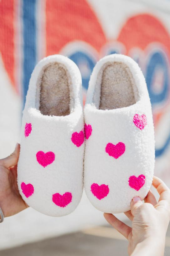 The Katydid Pink Hearts All Over Slippers For Women, fuzzy and white with bright pink heart patterns, are shown being held up against a colorful blurred background.