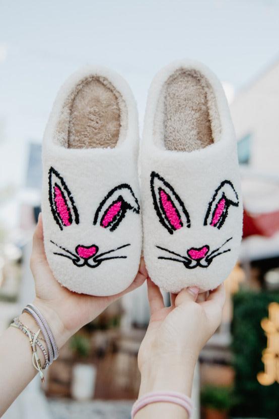 Hands hold up fluffy white slippers with cartoon bunny faces, featuring pink ears and noses, against a blurred outdoor background.