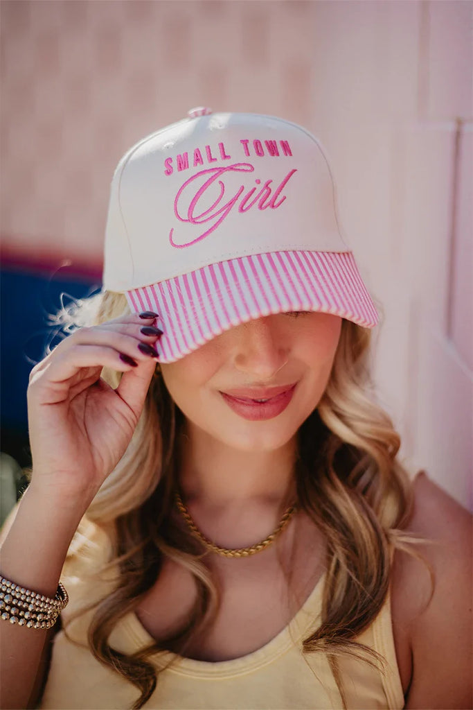 A woman with wavy blonde hair wears a Katydid Small Town Girl Vintage Trucker Hat for Women in cream and pink, smiling softly as she holds the brim. Shes styled in a yellow tank top and gold jewelry, radiating nostalgic, country-cute charm.