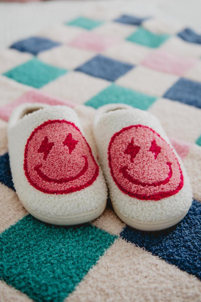 A pair of white slippers with pink lightning smiley faces on them sitting on a checkered floor