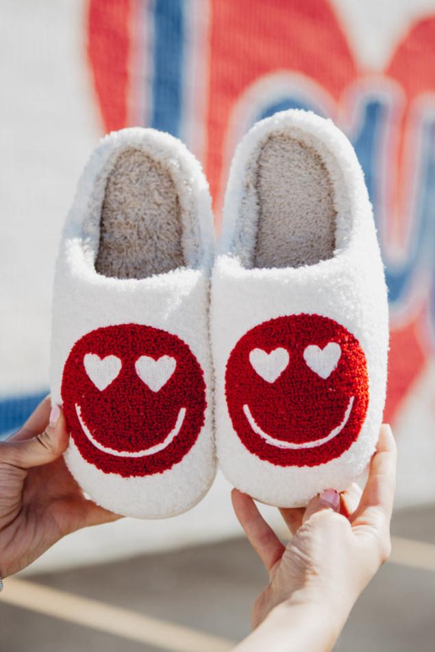 Two hands display a pair of soft, white Katydid slippers—fuzzy with red smiley faces and heart-shaped eyes—set against a blurred backdrop featuring bold, colorful text. These adorable slippers are part of the Build Your Own Katydid Set from Katydid.com 10 items for 60, a 350 value—perfect for anyone who loves cute accessories.
