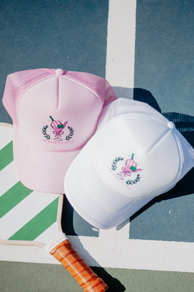Two trucker hats, one pink and one white, with pickleball graphic logos lie on a court. A green-striped pickleball paddle with an orange grip is nearby.