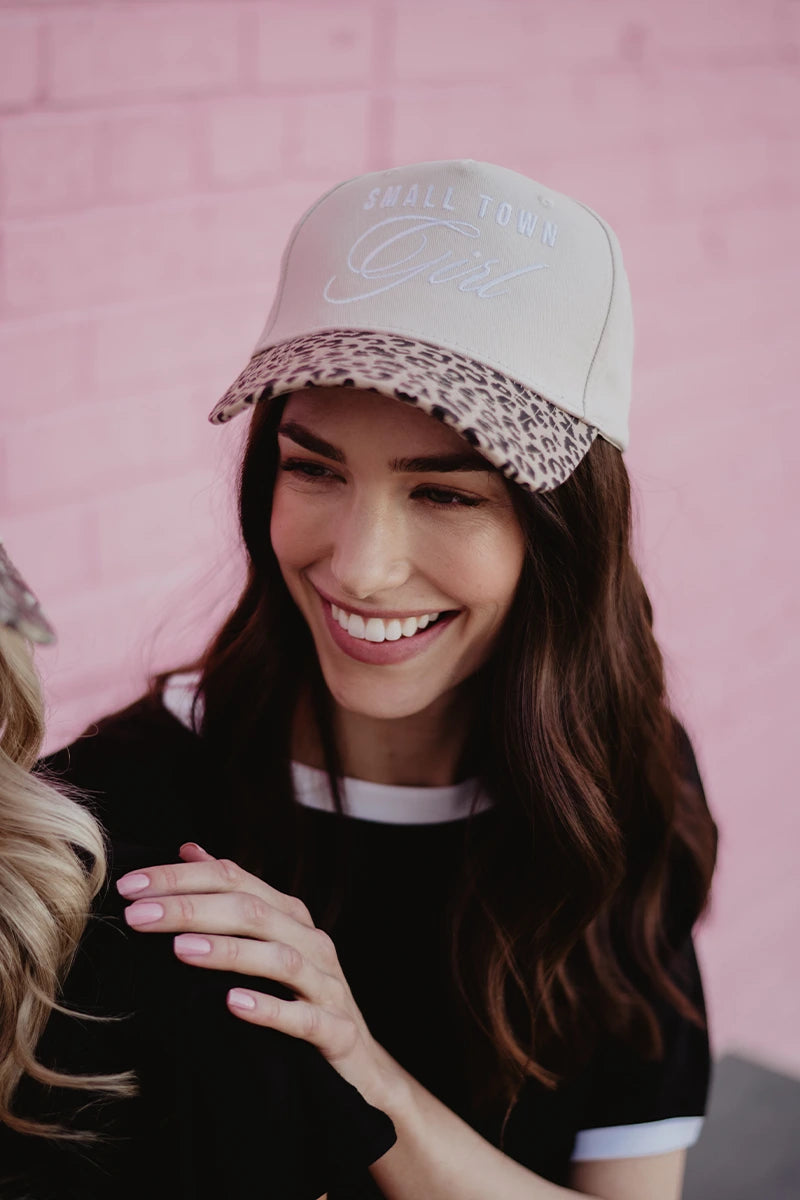 A smiling woman with long brown hair wears a Katydid Small Town Girl Vintage Trucker Hat—beige crown, leopard print brim—and a black tee with white trim, standing by a pink brick wall and showing off her country-cute style.