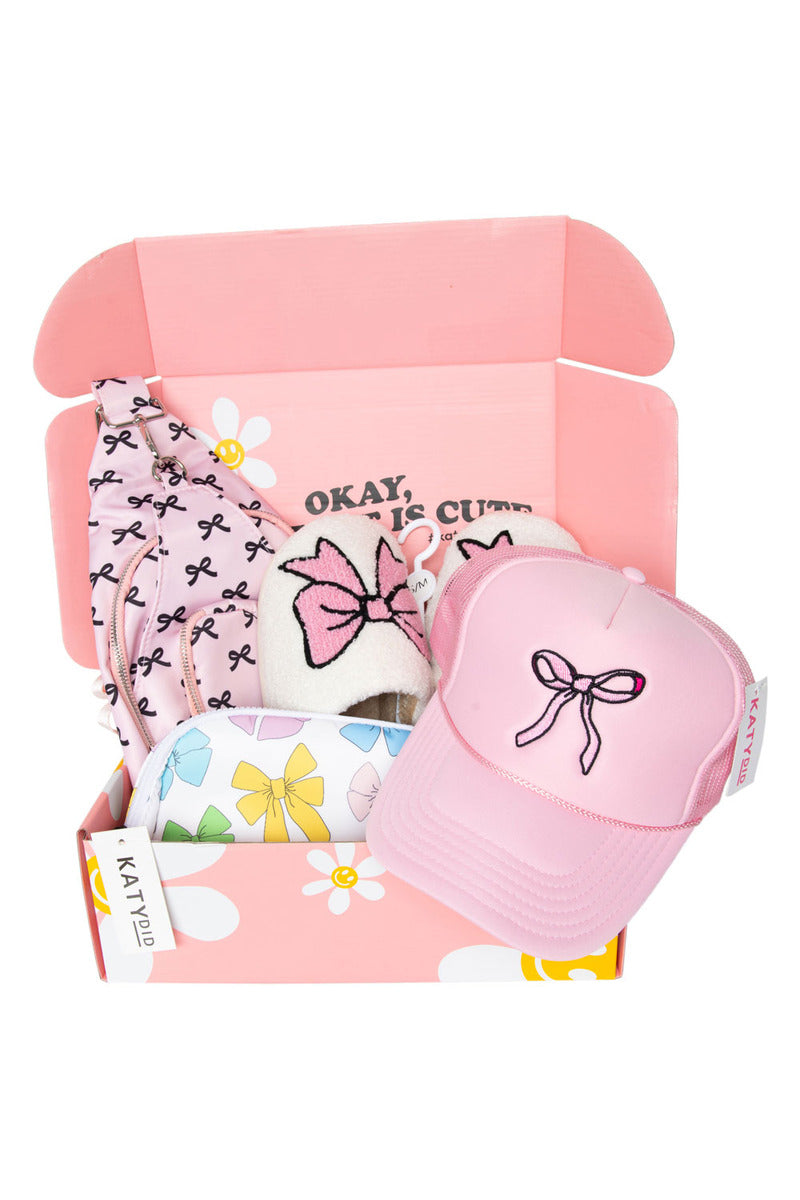 Pink gift box with bow-themed items: baby hat, bib, socks, pouch, and cap. Box reads "OKAY, THIS IS CUTE," set against a cheerful floral pattern.