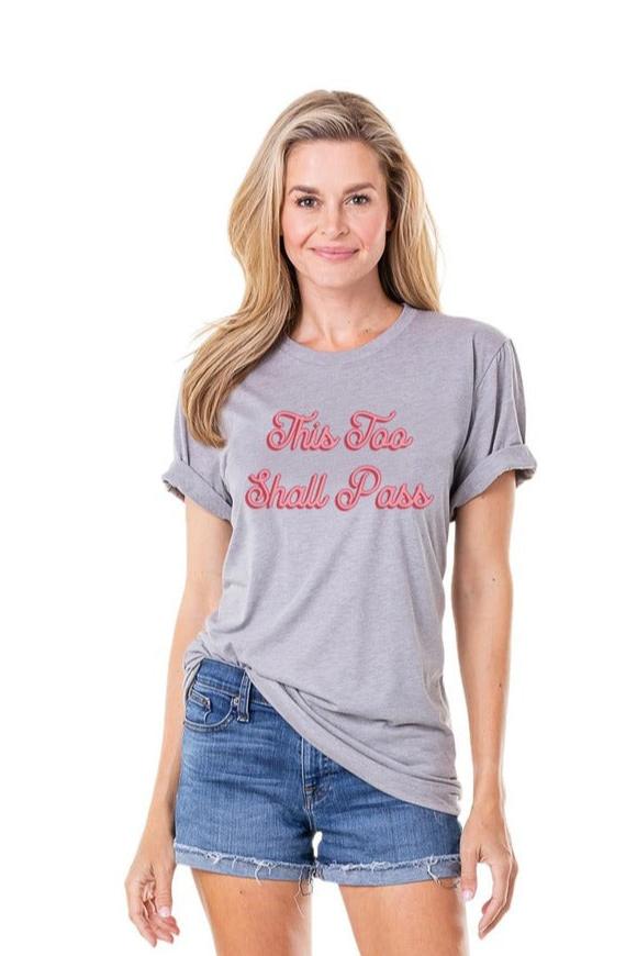 A woman in Katydid’s “This Too Shall Pass” Women’s T-Shirt—gray with pink cursive lettering—pairs it with denim shorts and smiles against a plain white background.