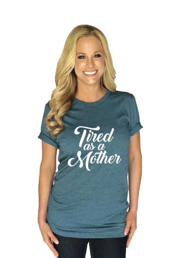 A smiling woman with long blonde hair models the Katydid “Tired As A Mother” T-Shirt, featuring a blue round neck design with white script, against a plain white background.