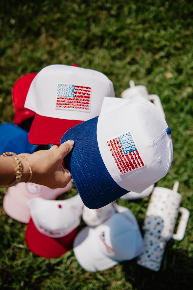 A hand holds a white baseball cap with a blue brim, featuring an American flag design. Other caps and a water bottle lie on green grass below.