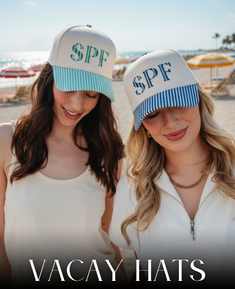 Two women on a sunny beach wear hats labeled SPF with blue and green striped brims. They smile, evoking a relaxed vacation vibe. Text reads, 'Vacay Hats.'