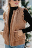 A woman stands outdoors in a cozy fall setting, wearing the Katydid Brown Garment Washed Quilted Ruffle Sleeve Vest over a black turtleneck and blue jeans, with sunglasses and her hand in her pocket amid blurred holiday décor.