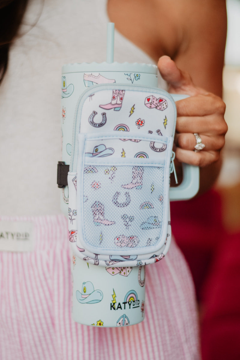 A person holds a pastel blue tumbler with a matching pouch adorned with playful cowboy boots, hats, and rainbows.