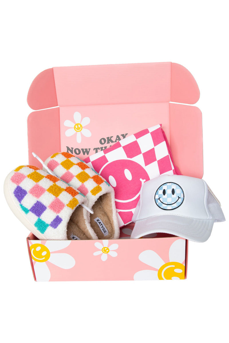 Open pink box with floral design containing checkered slippers, a pink checkered towel, and a white cap with a smiley face.