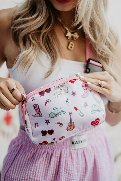 A woman with long blonde hair holds the Katydid Music Girl Icons Belt Bag, featuring pink patterns of cowboy hats, boots, hearts, guitars, and lips. She pairs it with a white top, pink skirt, layered necklaces, bright pink lipstick, and adjustable strap.