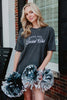 A smiling woman with long blonde hair holds silver and navy pom-poms outside. She wears a dark oversized Katydid Game Day Social Club Graphic Tee.