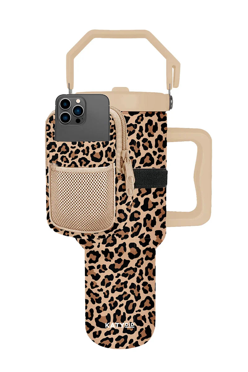 The Katydid Brown Leopard Tumbler Sling Bag holds your beige travel mug and features a handle, carrying strap, and a front pocket with mesh and zipper for your smartphone and secure storage.