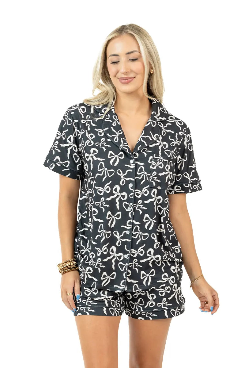A woman smiles in the Katydid Black Shorts Set Bow Pajamas—black short-sleeve button-up top and matching shorts with a white abstract bow print. She accessorizes with bracelets against a plain white background.