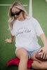 A woman with long blonde hair sits on a sports field, smiling as she looks down. She wears sunglasses, denim shorts, red cowboy boots, and the Katydid Game Day Social Club Graphic Tee while holding a football.