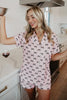 A smiling woman in a modern kitchen wears the Katydid Black  Pink Mini Coquette Bows Loungewear Set, holding a metal bowl and whisk. A glass of red wine rests on the white countertop beside her.