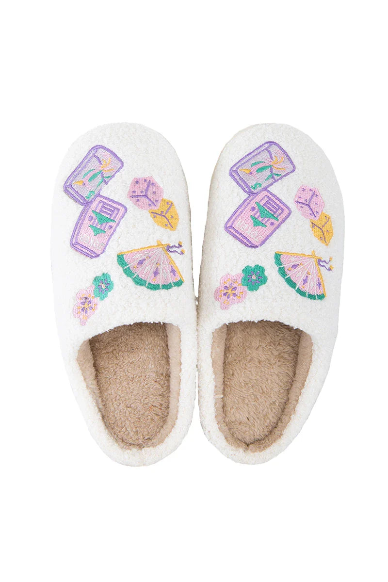 The Katydid Mahjong Pieces Cozy Slippers are white and fuzzy with tan interiors, featuring embroidered fans, dice, flowers, and pastel tiles for a soft and playful touch.
