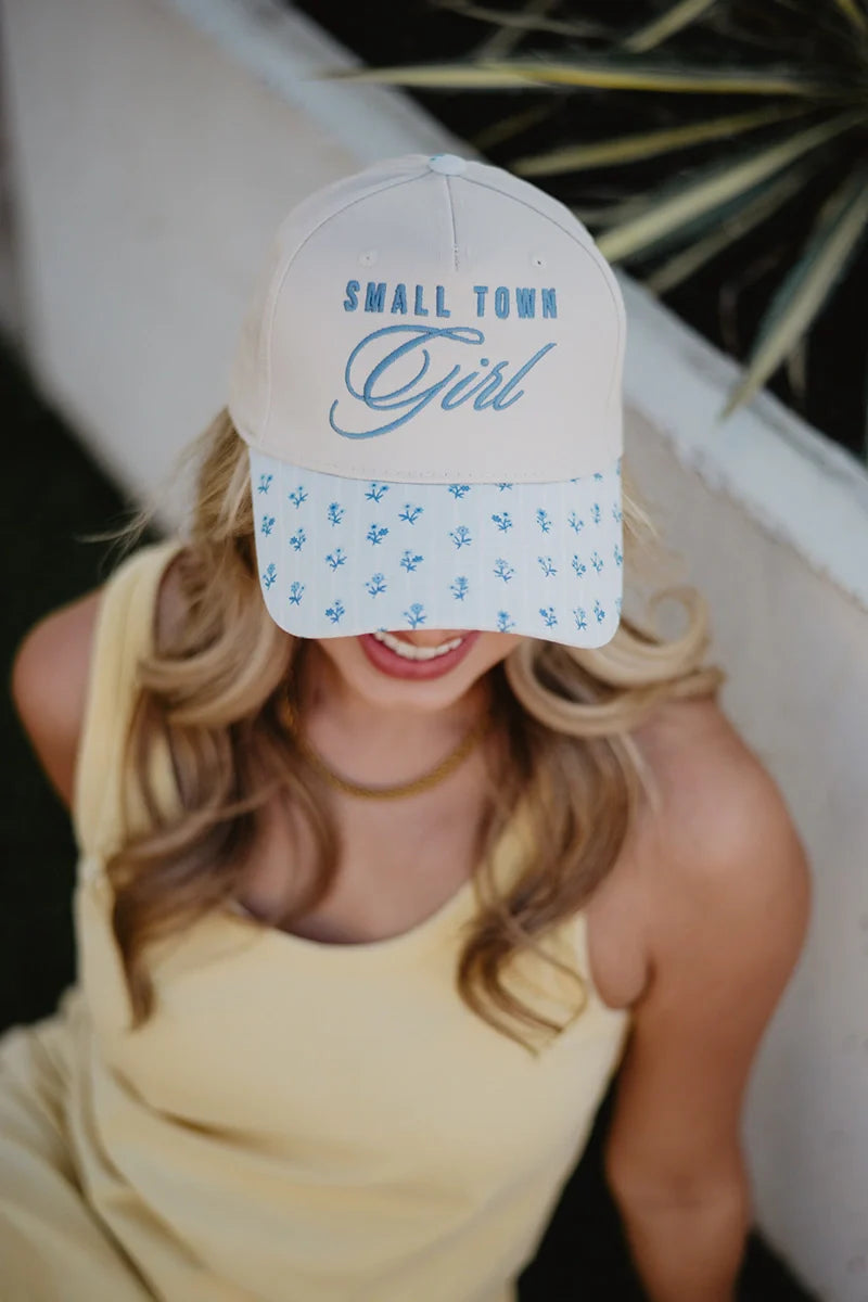 A woman in a yellow dress smiles outdoors, wearing the Katydid Small Town Girl Vintage Trucker Hat for Women with SMALL TOWN GIRL on the front and a blue floral brim. Her blonde hair cascades over her shoulders.