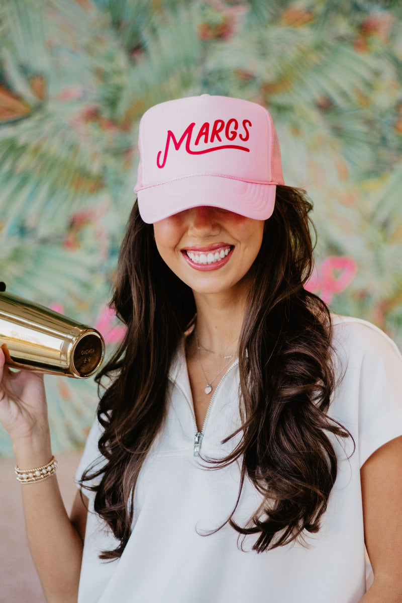 Smiling woman wearing a pink cap with "MARGS" text, holding a shiny cocktail shaker. Background features a colorful, tropical pattern.