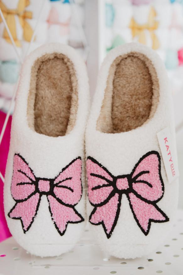 Cozy white slippers with pink bow designs displayed on a shelf.