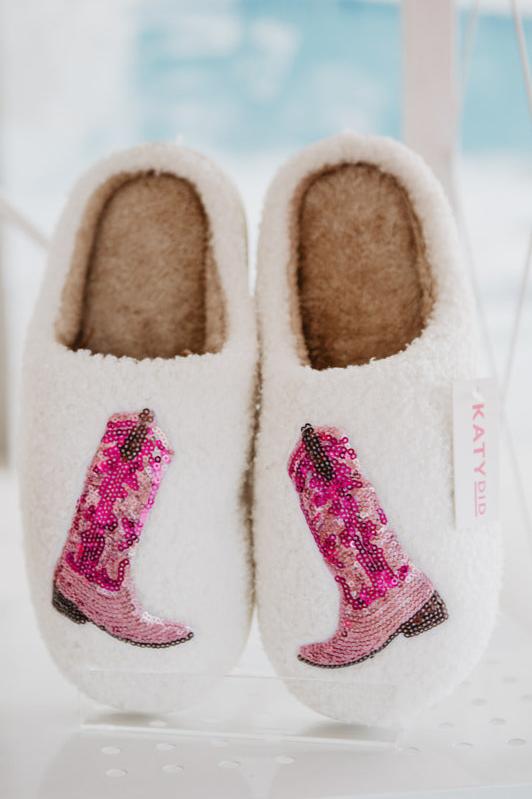 Cozy white slippers with a fluffy interior, adorned with sparkly pink cowboy boot designs.