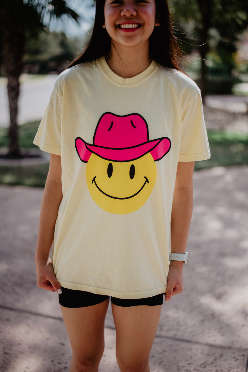 A person outdoors wears the Katydid Cowboy Happy Face Western Graphic Tee—light yellow with a big smiley face in a pink cowboy hat—paired with black shorts, smiling brightly with trees in the background.