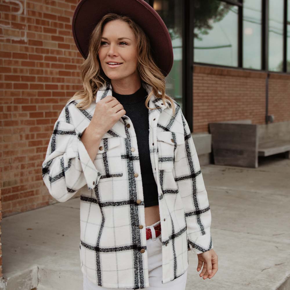 A woman smiles outdoors in the Katydid White and Black Plaid Shacket for Women, styled with a wide-brimmed hat and black crop top—an inspiring look for chic fall outerwear.