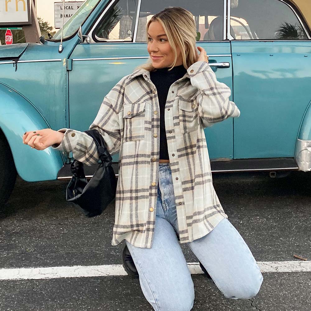 A woman kneels beside a vintage blue car, smiling as she touches her hair, embracing fall in the Katydid Plaid Shacket for Women over a black turtleneck and light blue jeans, with a small black handbag.