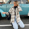 A woman kneels beside a vintage blue car, smiling as she touches her hair, embracing fall in the Katydid Plaid Shacket for Women over a black turtleneck and light blue jeans, with a small black handbag.