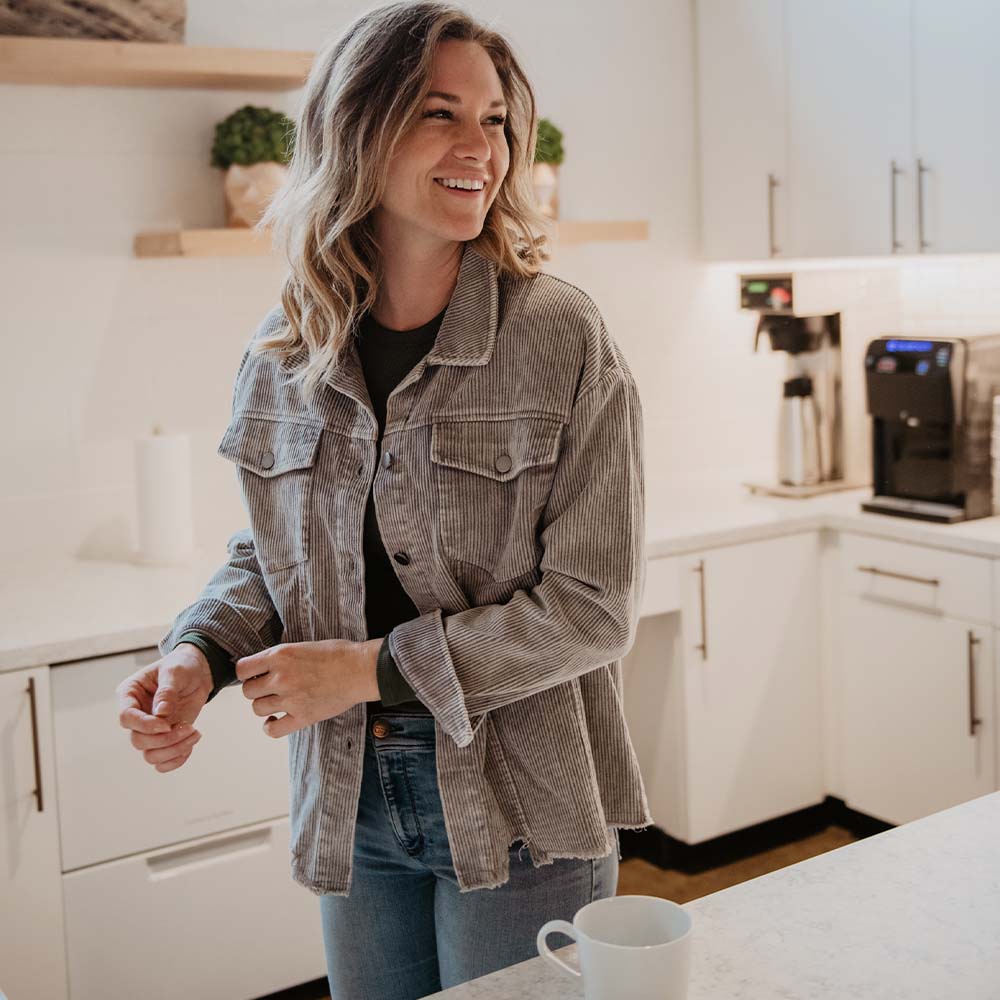A woman with wavy hair smiles in a bright kitchen, wearing Katydids Neutral Corduroy Distressed Shacket for Women and jeans. A white mug sits on the counter in front of her.