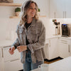 A woman with wavy hair smiles in a bright kitchen, wearing Katydids Neutral Corduroy Distressed Shacket for Women and jeans. A white mug sits on the counter in front of her.